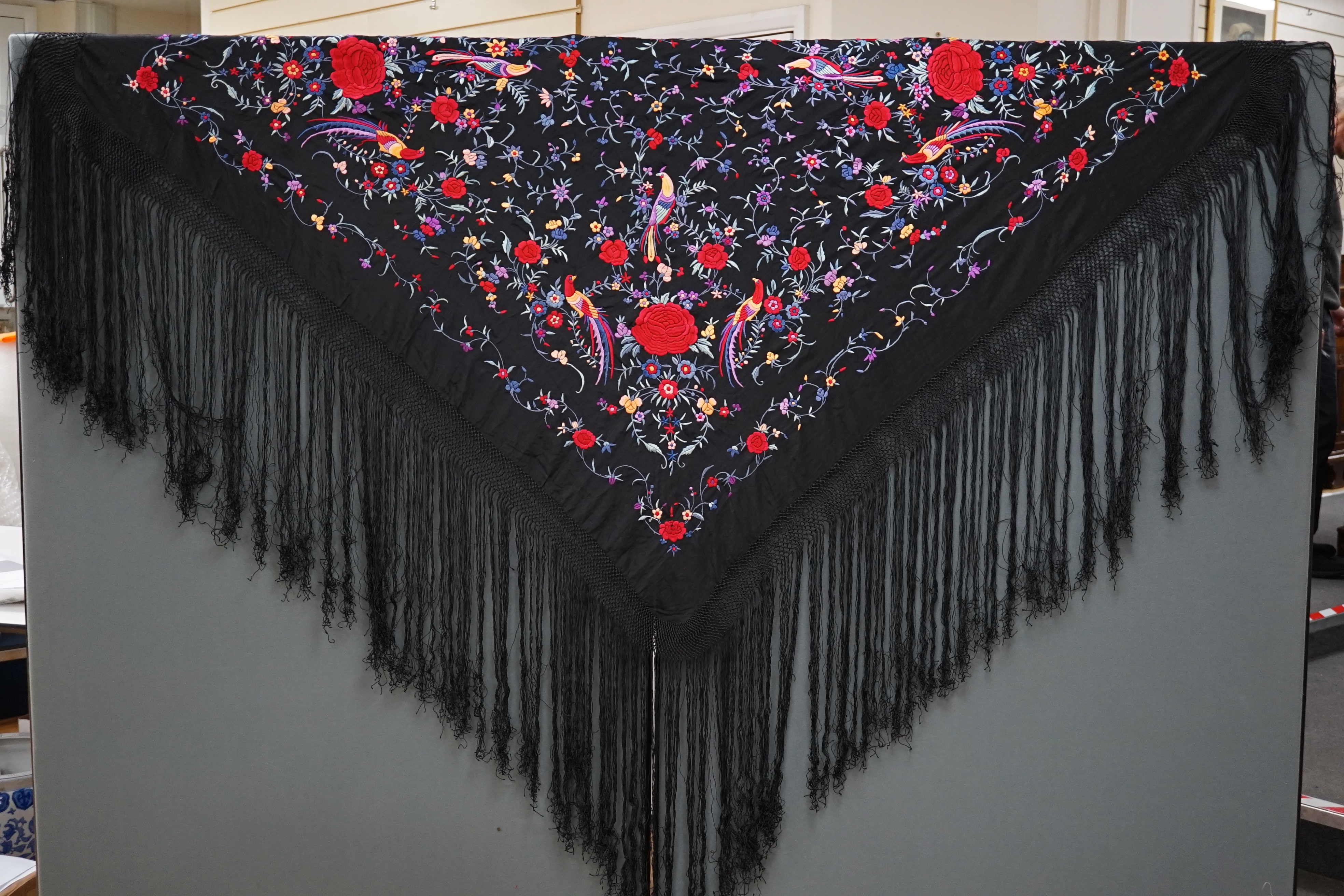 An early 20th century Chinese black silk fringed shawl with polychrome silk embroidery, embroidered with meandering floral design, the corners embroidered with three brightly coloured birds of paradise, large red roses a